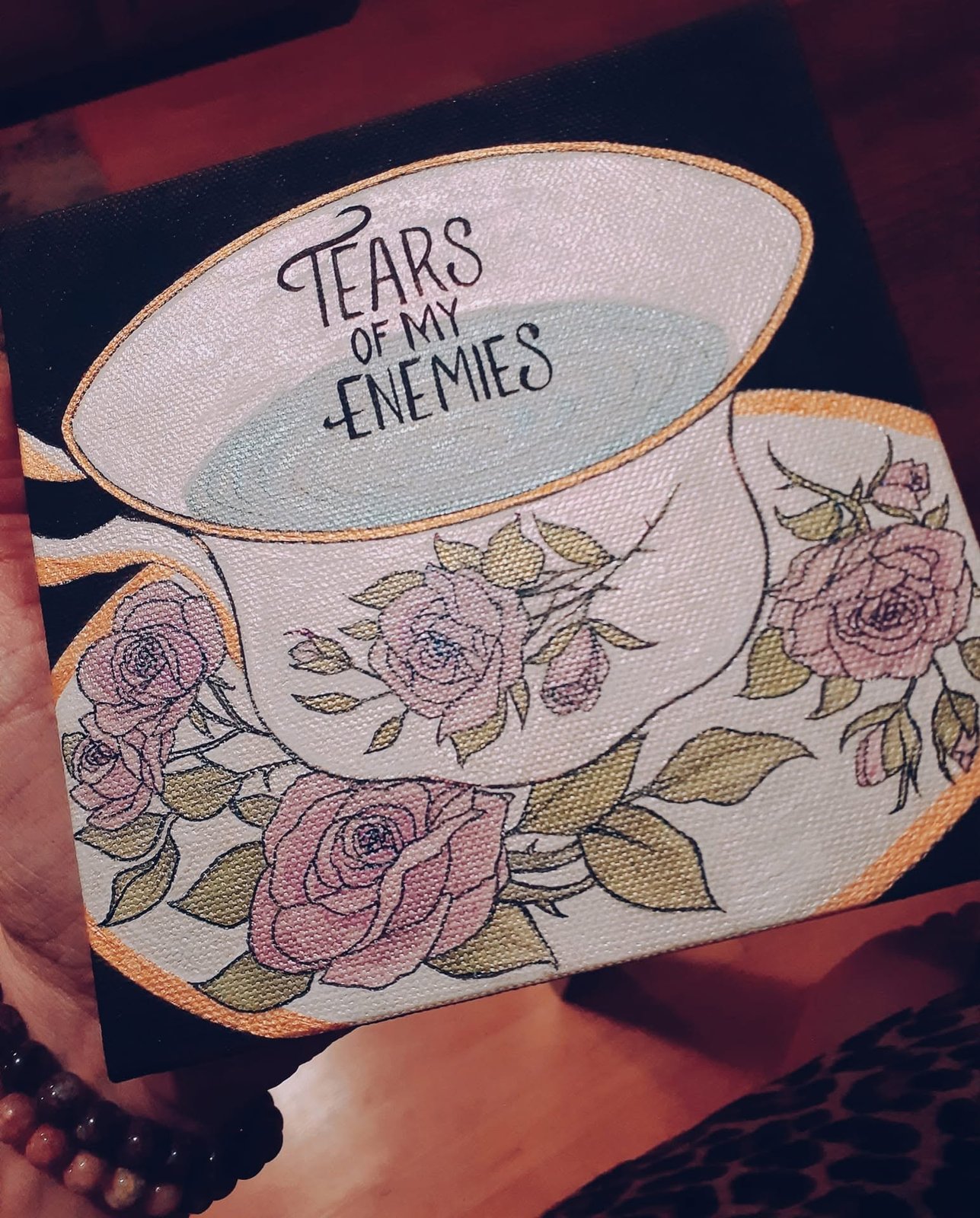 Acrylic painting of a rose-patterned teacup and saucer lettered 'Tears of my enemies', held up by the artist