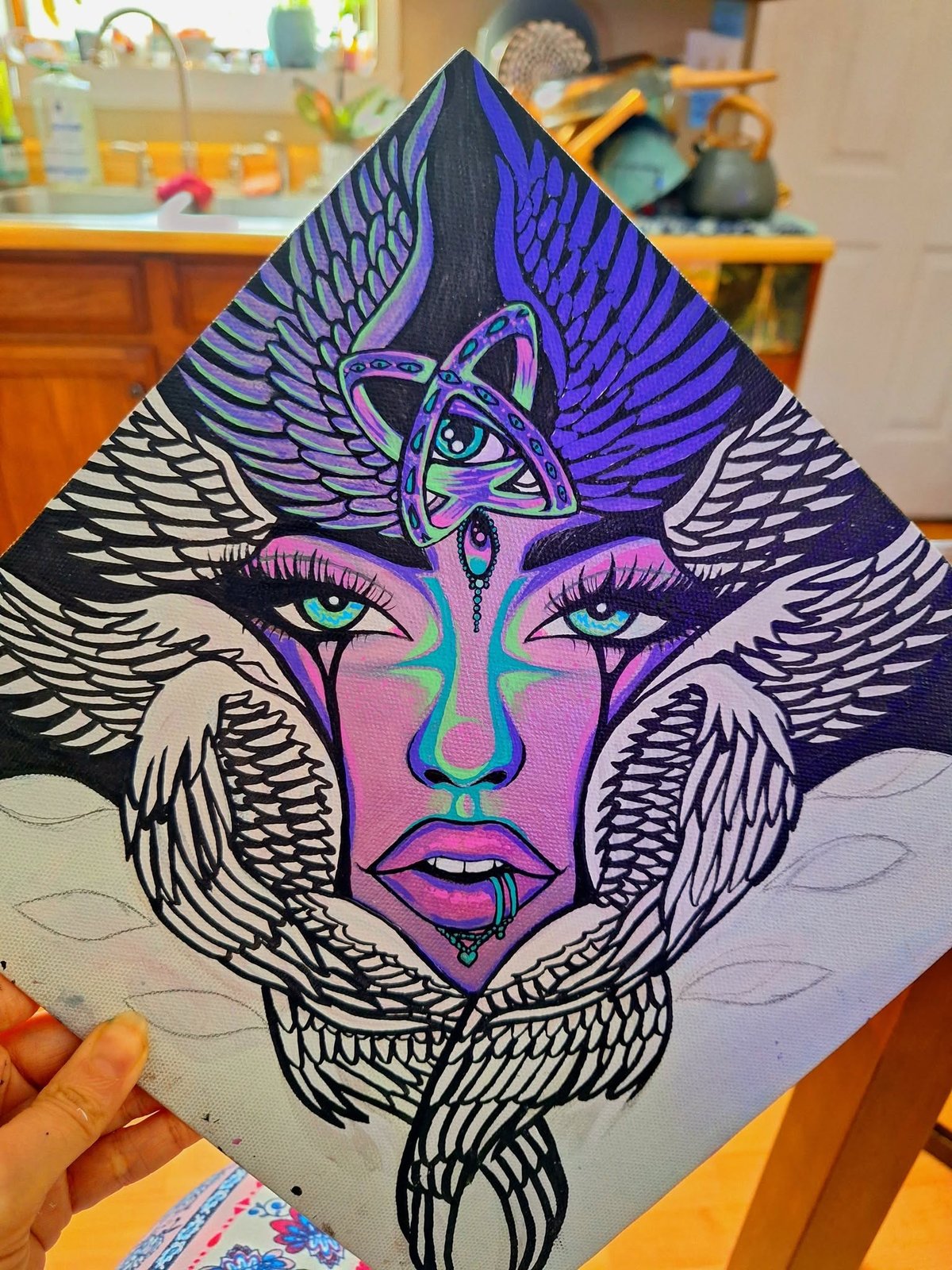 Painting on a diamond-shaped board of a violet-skinned face with a third eye inside a triquetra knot, framed by black and white feathered wings