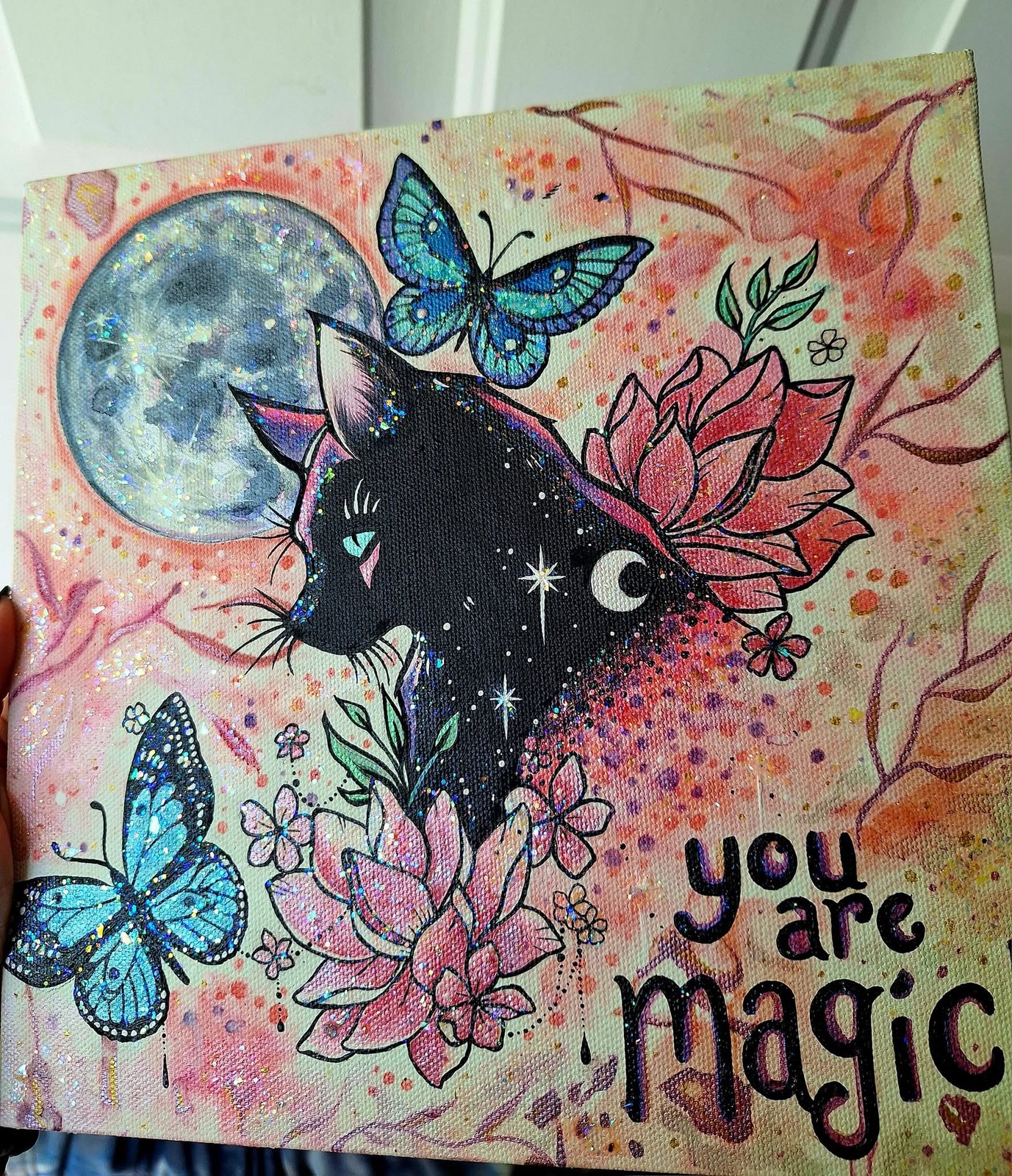 Painting of a black cat with a crescent mark on its chest beneath a full moon, blue butterflies and pink lotus flowers around it, lettered 'you are magic'
